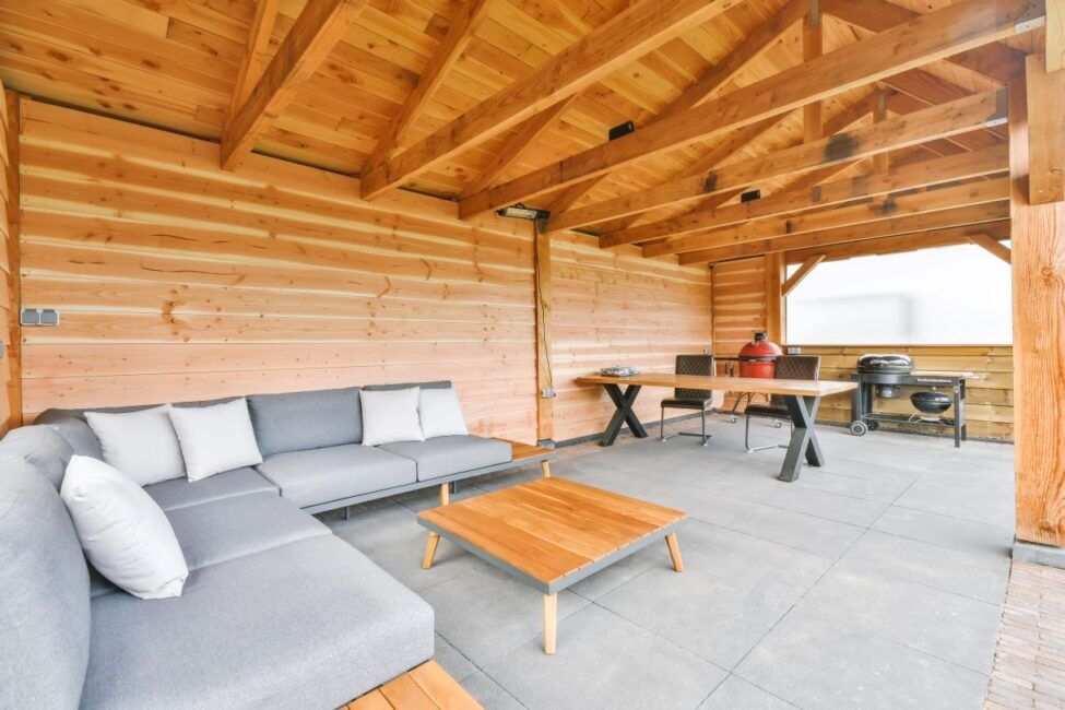 Дървен навес с луксозно ъглово канапе, голяма маса за хранене и барбекю на циментови плочи