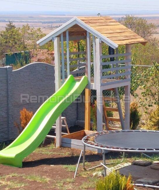 Детска площадка с дървена конструкция, пързалка, люлка и пясъчник, разположена на фона на красив пейзаж.